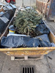 A picture of a skip containing black bag rubbish, paint tins and a real christmas tree.